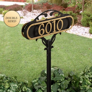 May include: A double-sided house number sign featuring the digits 6010 in gold metal mounted on a black oval frame with decorative scrollwork. The sign is attached to a vertical black metal post, designed for outdoor display in a garden setting.