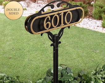 Puede incluir: Un letrero de número de casa de doble cara con los dígitos 6010 en metal dorado, montado sobre un marco ovalado negro con adornos decorativos. El letrero está sujeto a un poste vertical de metal negro para su exhibición en exteriores.