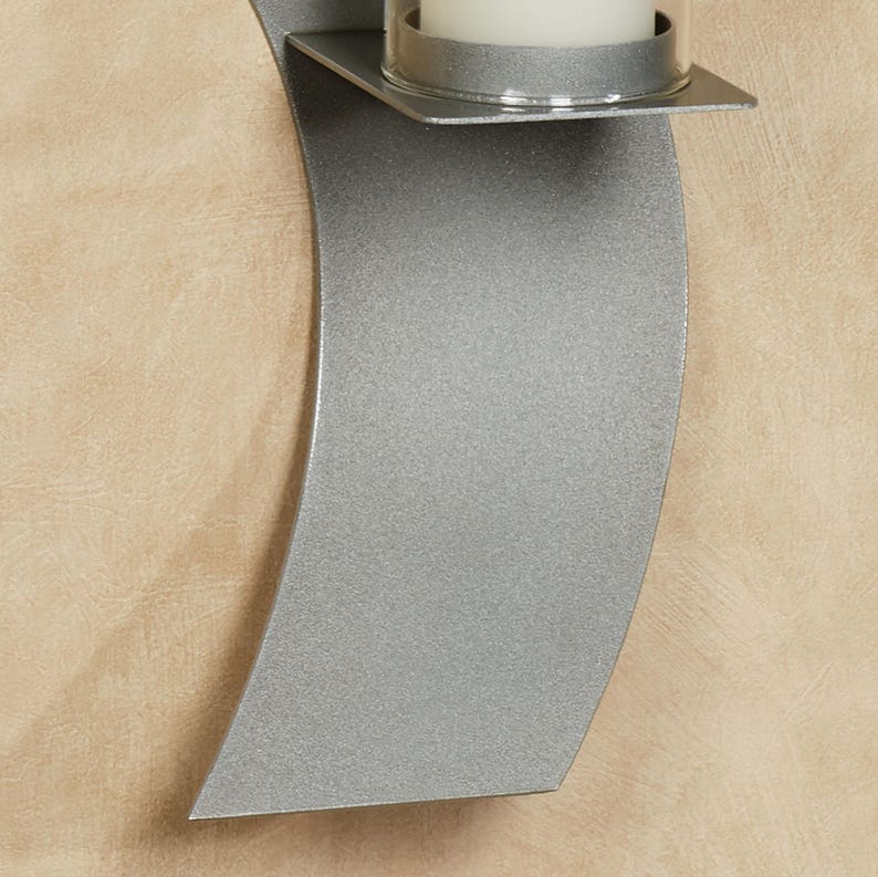 Puede incluir: Un aplique de pared para velas con una placa trasera de metal plateado curvado y acabado texturizado. En la parte superior, un peque&ntilde;o estante met&aacute;lico plano sostiene un soporte cil&iacute;ndrico de vidrio transparente con una vela blanca.