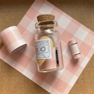 May include: A small glass bottle with a cork stopper, containing a heart-shaped piece of paper with a cassette tape design and the text "Tape in a Bottle". The bottle is sitting on a pink and white gingham patterned surface.