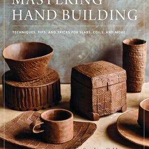 May include: A book cover titled "Mastering Hand Building" with various handmade clay objects. The objects include a bowl, a box, a cup and saucer, and a tall cylinder. The objects are brown and have textured surfaces.