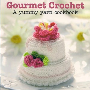 Könnte beinhalten: Ein Buchcover mit dem Titel Gourmet Crochet: A yummy yarn cookbook, das eine zweistöckige, weiß gehäkelte Torte zeigt. Die Torte ist mit einer großen rosa gehäkelten Blume oben und kleineren rosa Blüten mit grünen Blättern an der Seite verziert.