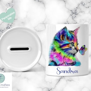 May include: White ceramic money box with a slot and a matching mug. The mug features a colourful illustration of a kitten with a butterfly and the name "Sandra" in blue script. The background is a marble pattern.