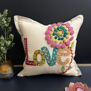 May include: A beige pillow with the word "LOVE" embroidered in colorful fabric. The letters are decorated with green leaves and a large pink flower with yellow and green polka dots.