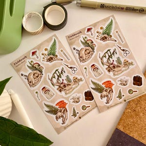 May include: Two sticker sheets with illustrations of animal skulls, mushrooms, ferns, and other botanical elements. The stickers are labeled "Fieldwork" and feature a variety of detailed designs in shades of brown, green, and orange. Other stationery items are in the background.