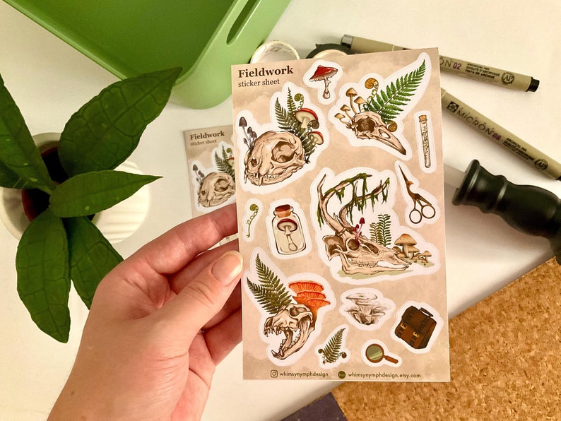 May include: A sticker sheet with a beige background featuring illustrations of animal skulls, mushrooms, ferns, and other botanical elements. The text "Fieldwork sticker sheet" is printed on the sheet. The stickers have a vintage, nature-inspired aesthetic.