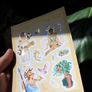 May include: A sheet of gold foil stickers featuring illustrations of Greek goddesses. The stickers include images of Persephone, Medusa, Athena, and Artemis. The sheet is decorated with a gold Greek key border.