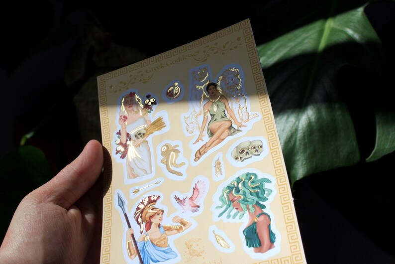 May include: A sheet of gold foil stickers featuring illustrations of Greek goddesses. The stickers include images of Persephone, Medusa, Athena, and Artemis. The sheet is decorated with a gold Greek key border.