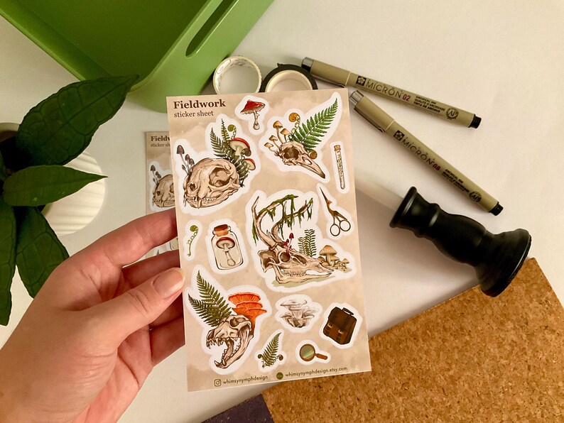 May include: A sticker sheet with the text "Fieldwork" featuring illustrations of skulls, mushrooms, ferns, and other botanical elements. The stickers are in shades of brown, green, and beige. Two Micron pens, a black stamp, and washi tape are also visible.