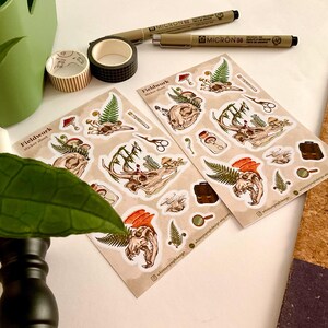 May include: Two sticker sheets with botanical and skull illustrations, along with pens, washi tape, and a green container. The stickers feature detailed drawings of skulls, ferns, and mushrooms. The text "Fieldwork sticker sheet" is visible on the stickers.
