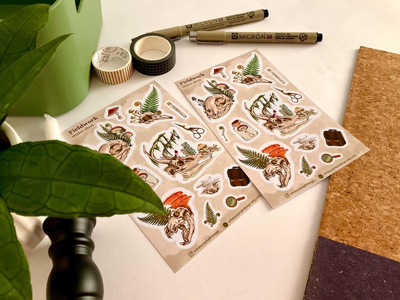 May include: Two sticker sheets with botanical and skull illustrations, along with pens, washi tape, and a green container. The stickers feature detailed drawings of skulls, ferns, and mushrooms. The text "Fieldwork sticker sheet" is visible on the stickers.