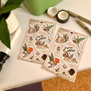 May include: Two sticker sheets with a beige background featuring skull and antler illustrations, mushrooms, ferns, and other botanical elements. The stickers are accompanied by pens, washi tape, and a plant in a white pot. The sticker sheet is labeled "Fieldwork".