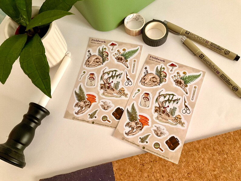 May include: Two sticker sheets with a beige background featuring skull and antler illustrations, mushrooms, ferns, and other botanical elements. The stickers are accompanied by pens, washi tape, and a plant in a white pot. The sticker sheet is labeled "Fieldwork".