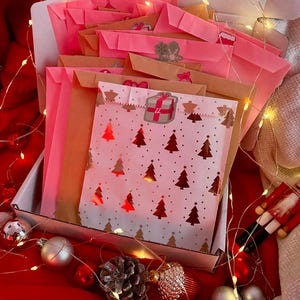 Peut inclure: Une boîte en carton blanc remplie de plusieurs petites enveloppes en papier dans des tons rose et marron. L'enveloppe avant présente un fond blanc avec un motif de sapins de Noël rouge métallisé et un autocollant avec le numéro 22.