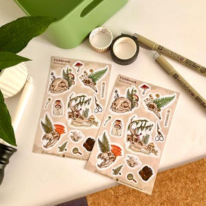 May include: Two sticker sheets with a Fieldwork theme, featuring illustrations of skulls, mushrooms, ferns, and other natural elements. Also visible are two Micron 02 pens, washi tape, and a green container. The stickers have a vintage aesthetic.