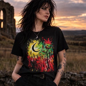 Op de afbeelding: Een zwart t-shirt met korte mouwen en een levendige grafische print op de voorzijde. Het ontwerp toont een gele maansikkel, zwarte krullende rankpatronen en een abstracte achtergrond met verfspatten in de kleuren rood, groen en geel.