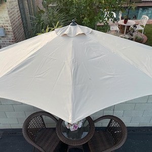 May include: A large, cream-colored patio umbrella with a scalloped edge. The umbrella is open, providing shade over a small glass-topped table and two wicker chairs. The scene is set outdoors.