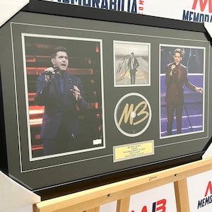 Puede incluir: Una foto en blanco y negro enmarcada de un hombre cantando en un escenario. La foto está rodeada de un marco negro con un borde dorado. El marco está sobre un caballete de madera. La foto está firmada por el cantante. La foto se titula "Michael Bublé Higher" y la fecha "Released March 25, 2022" está impresa debajo del título. La foto también está etiquetada como "Hand Signed CD Block".