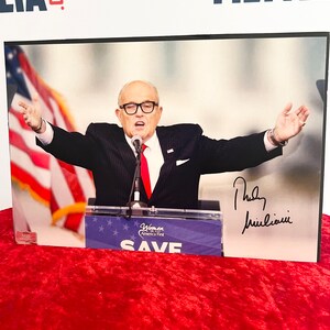 Könnte beinhalten: Ein gerahmtes Foto eines Mannes im Anzug, der an einem Podium spricht. Er trägt eine rote Krawatte und ein Mikrofon steht vor ihm. Das Podium trägt ein Schild mit der Aufschrift "Women America First SAVE". Der Mann unterschreibt seinen Namen "Rudy Giuliani" mit schwarzer Tinte.