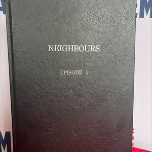 May include: Black leather-bound book with the title "Neighbours" and "Episode 1" printed in silver lettering.