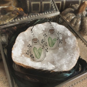 Op de afbeelding: Een paar oorbellen met glazen flesjes gevuld met groene kristallen. De flesjes zijn omringd door delicate bloemmotieven en zijn geplaatst op een witte achtergrond.