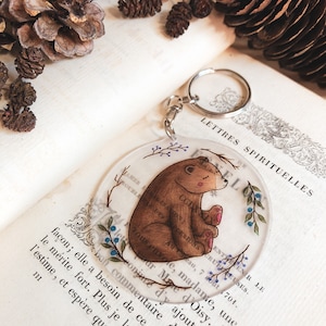 May include: A round, clear keychain with a brown bear illustration. The bear is sitting with its paws tucked in and has a pink nose and blush on its cheeks. The background of the keychain is a light blue with white branches and purple berries.