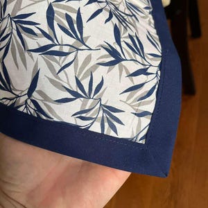 Op de afbeelding: Een hoek van een witte stof met een patroon van blauwe en grijze bladeren, omlijst door een marineblauwe rand. De stof wordt vastgehouden, wat het ontwerp en het kleurcontrast benadrukt.