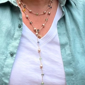 May include: A multi-strand lariat necklace featuring a delicate gold-toned chain interspersed with small, round, matte-finish beads in shades of cream, pale green, and dark blue, worn over a white t-shirt and a light green button-down shirt.