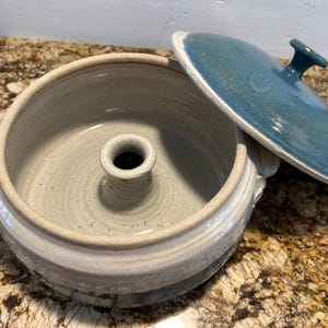 May include: A round ceramic baking dish with a light gray interior and a central chimney column. The exterior features a white and blue glaze with two side handles. A matching circular lid with a blue glaze and a small knob sits to the side.