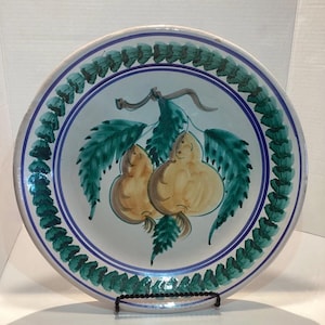 May include: A decorative ceramic plate featuring a hand-painted design of two yellow pears with green leaves. The plate has a white background with a blue and green border. The plate is displayed on a black metal stand.