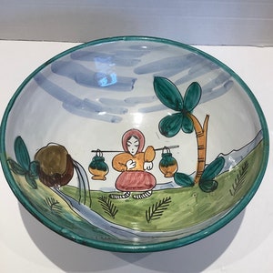 May include: A large, white ceramic bowl with a hand-painted design. The bowl features a woman carrying jugs on a stick, a palm tree, and a stream. The bowl has a green rim and a blue and white sky.
