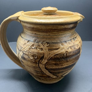 May include: A handmade ceramic pitcher with a lid. The pitcher is tan and brown with a tree branch design. It has a handle and a lid with a small knob. The pitcher is approximately 7 inches tall.