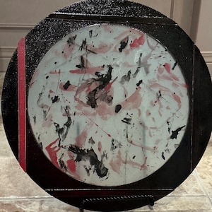 Könnte beinhalten: Runder dekorativer Teller mit schwarzem Rahmen und einem zentralen Design mit abstrakten Spritzern in Rosa, Schwarz und Weiß. Ein vertikaler roter Streifen akzentuiert die Seite. Der Teller wird auf einem schwarzen Metallständer präsentiert.