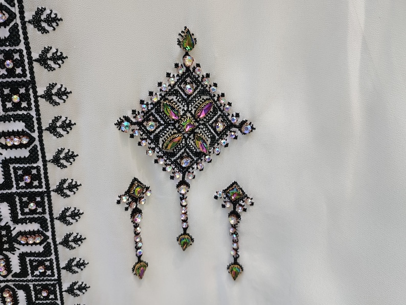 May include: A white fabric with black embroidery and iridescent rhinestones in a geometric pattern. The design features a large diamond shape with three smaller diamond shapes hanging below.