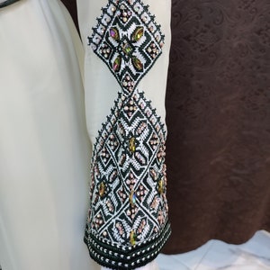 May include: A white dress with intricate black and green embroidery and embellishments. The embroidery features a geometric pattern with small, iridescent beads.