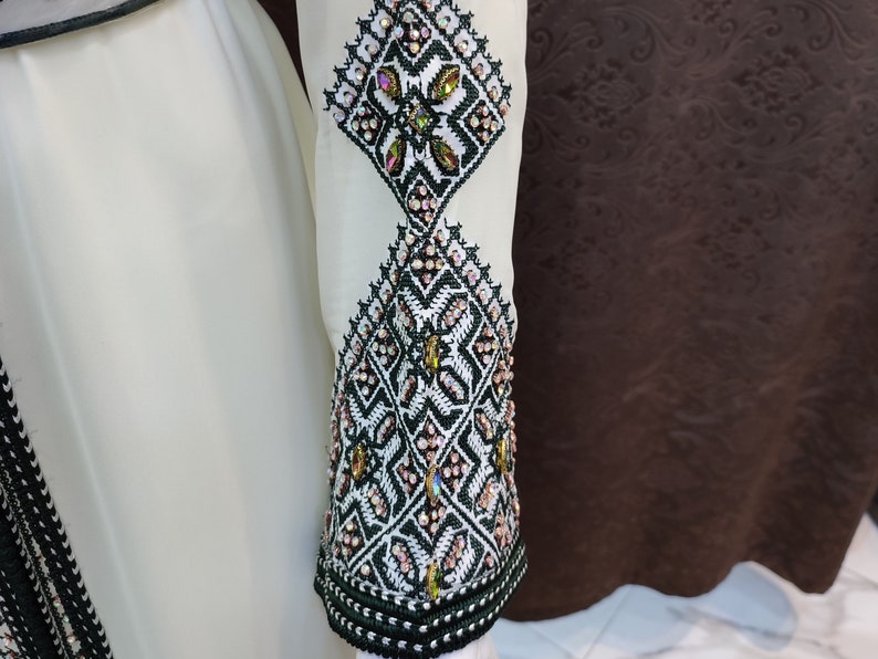 May include: A white dress with intricate black and green embroidery and embellishments. The embroidery features a geometric pattern with small, iridescent beads.
