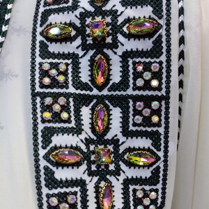 May include: A white fabric with a green and white embroidered trim. The trim features a geometric pattern with iridescent rhinestones.