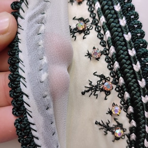 May include: Close-up of a white fabric with green and white trim. The fabric is decorated with black embroidery and iridescent rhinestones.