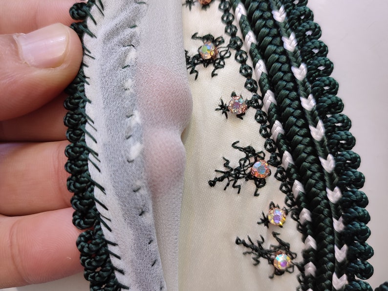 May include: Close-up of a white fabric with green and white trim. The fabric is decorated with black embroidery and iridescent rhinestones.