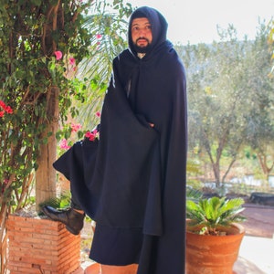 May include: A man wearing a dark blue hooded cloak standing in front of a brick wall and green plants.