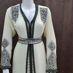 May include: A white long-sleeved dress with black and green trim and intricate embroidery. The dress has a V-neckline and a belt.