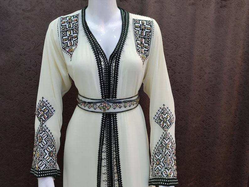 May include: A white long-sleeved dress with black and green trim and intricate embroidery. The dress has a V-neckline and a belt.