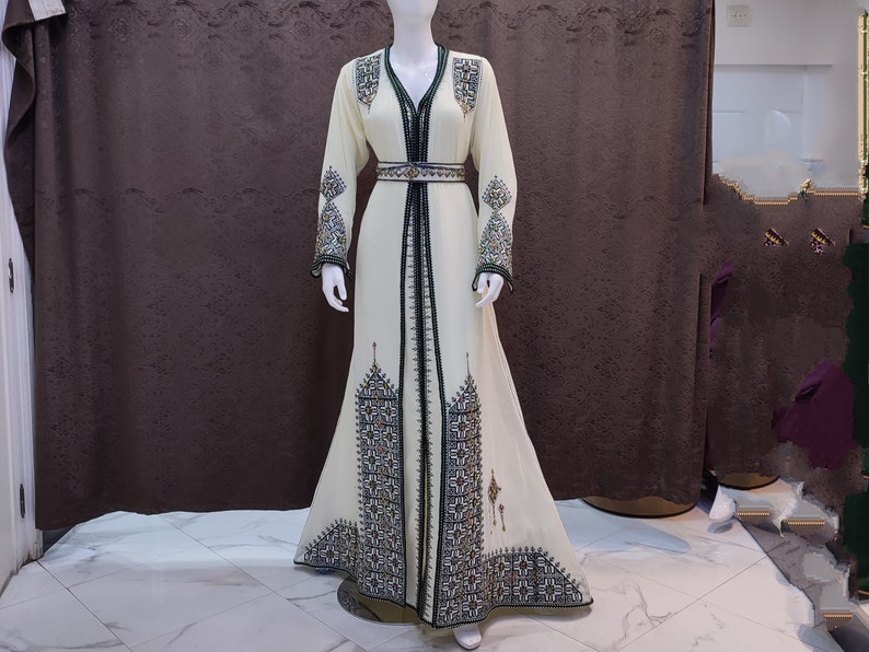 May include: A long, white dress with intricate black and green embroidery. The dress has a V-neckline and a belt at the waist.