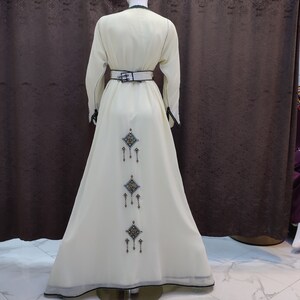 May include: A white maxi dress with a black belt and intricate embroidery on the back. The embroidery features a repeating pattern of diamonds with dangling beads.