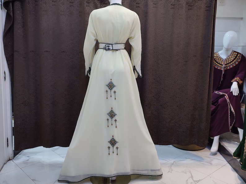 May include: A white maxi dress with a black belt and intricate embroidery on the back. The embroidery features a repeating pattern of diamonds with dangling beads.