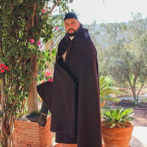 May include: A man wearing a brown hooded cloak stands in front of a brick wall and a potted plant. He is wearing a black hat and black shoes.