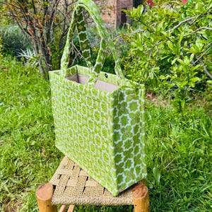 Peut inclure: Un sac fourre-tout vert citron avec un motif géométrique de formes blanches et beiges. Le sac a des poignées assorties et est posé sur un petit tabouret en bois avec une assise tissée. Le fond est un jardin verdoyant.