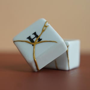 May include: Two white square ceramic tiles featuring a black letter H on the front surface. The tiles are repaired with gold-colored lines that mimic the kintsugi technique, creating a distinct pattern across the white surfaces of the objects.