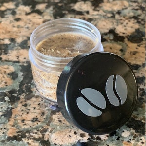 May include: A small clear jar with a black lid containing a brown coffee scrub. The lid has two grey coffee bean icons.