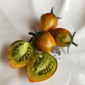 Puede incluir: Primer plano de cuatro tomates verdes y naranjas cortados por la mitad y enteros en un plato blanco. Los tomates tienen un patrón rayado y son una variedad llamada 'Green Zebra'.